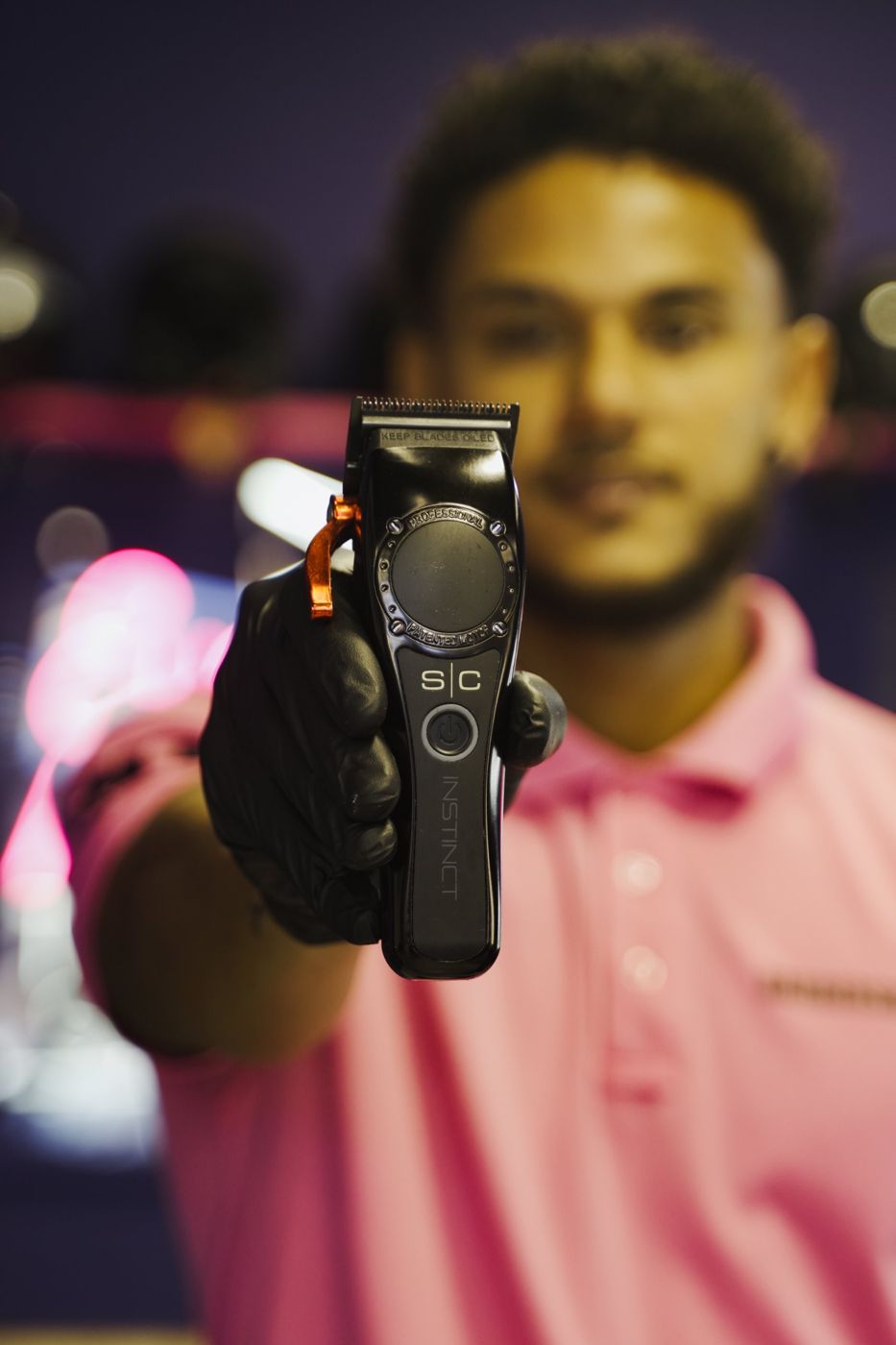 Barber in a pink polo holding clippers toward the camera, face out of focus